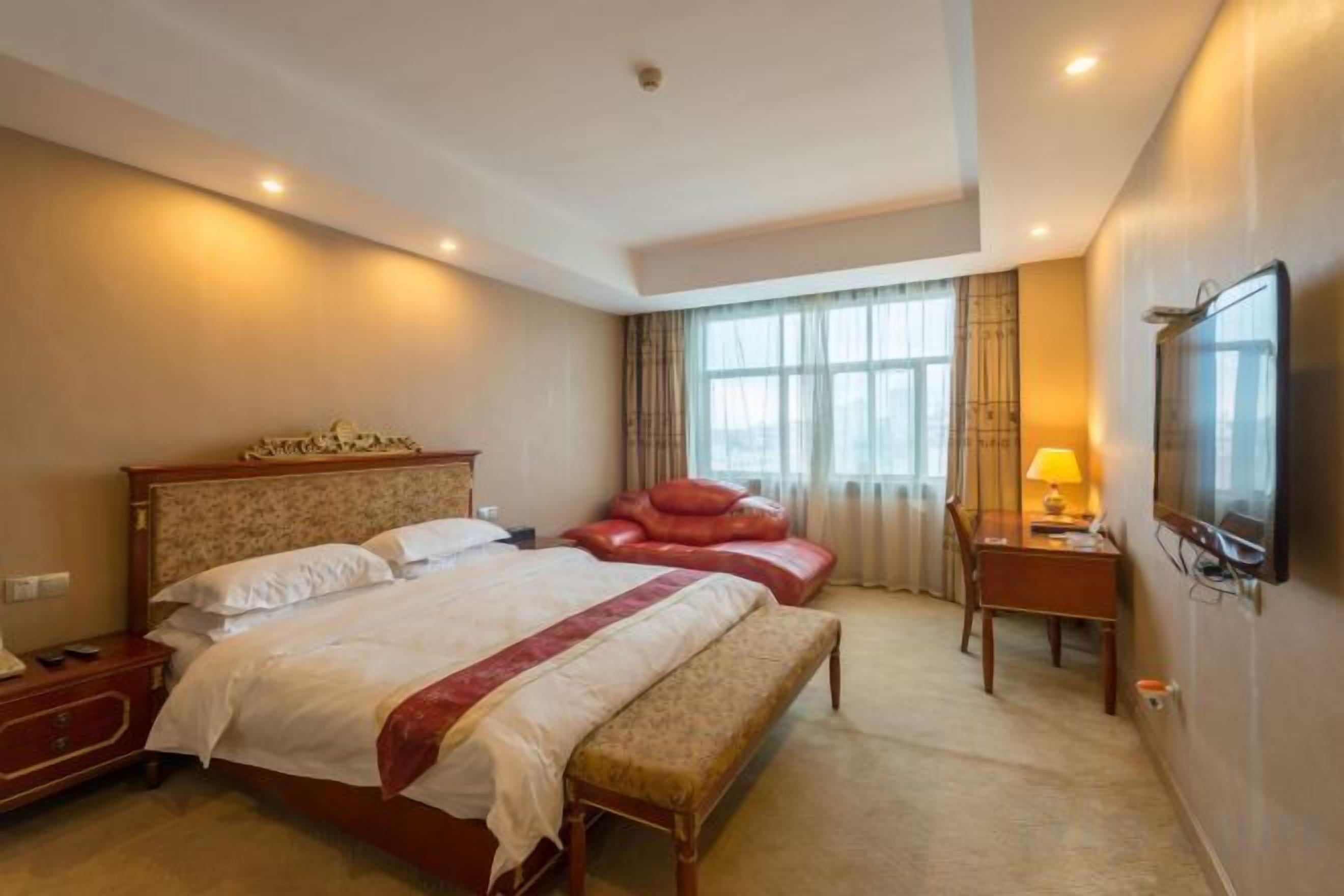 Een hotelkamer met een groot bed, een bankje, een bureau met lamp en een aan de muur bevestigde televisie.