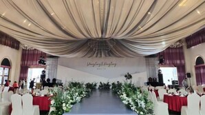 Une salle de banquet avec une longue table dressée pour un événement formel, recouverte d’un tissu blanc et entourée de chaises blanches.