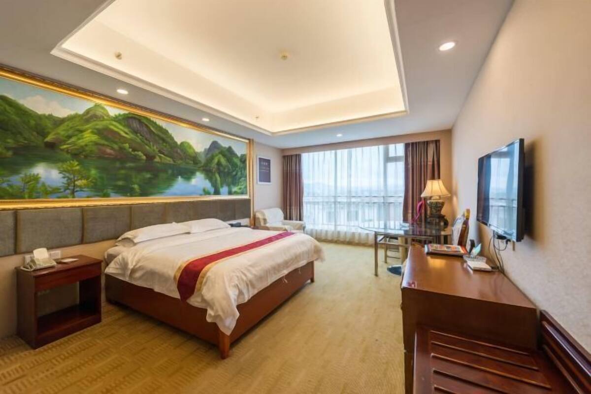A hotel room with a bed, a desk, a chair, a TV, and a large window with curtains.