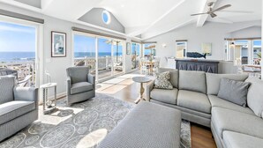 A spacious living area with a large sofa, a patterned rug, and a dining table. There is a balcony with a view of the ocean.