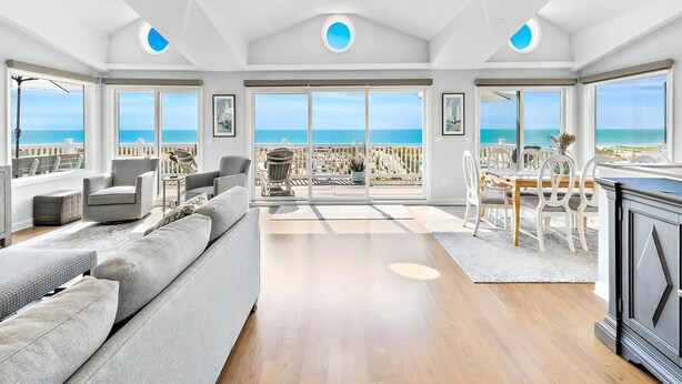 A spacious living area with a large sofa, a dining table with chairs, and a kitchen area. The room has large windows offering a view of the beach and ocean.