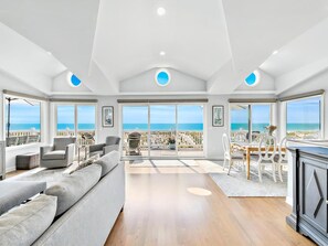 Amplia sala de estar con un sofá grande, una mesa de comedor con sillas y una zona de cocina. La habitación tiene ventanas amplias con vistas a la playa y al océano.