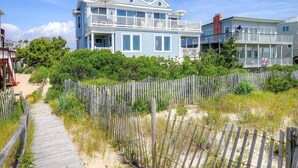 A two-story beachfront house with a wooden deck and a sandy path leading to the entrance.