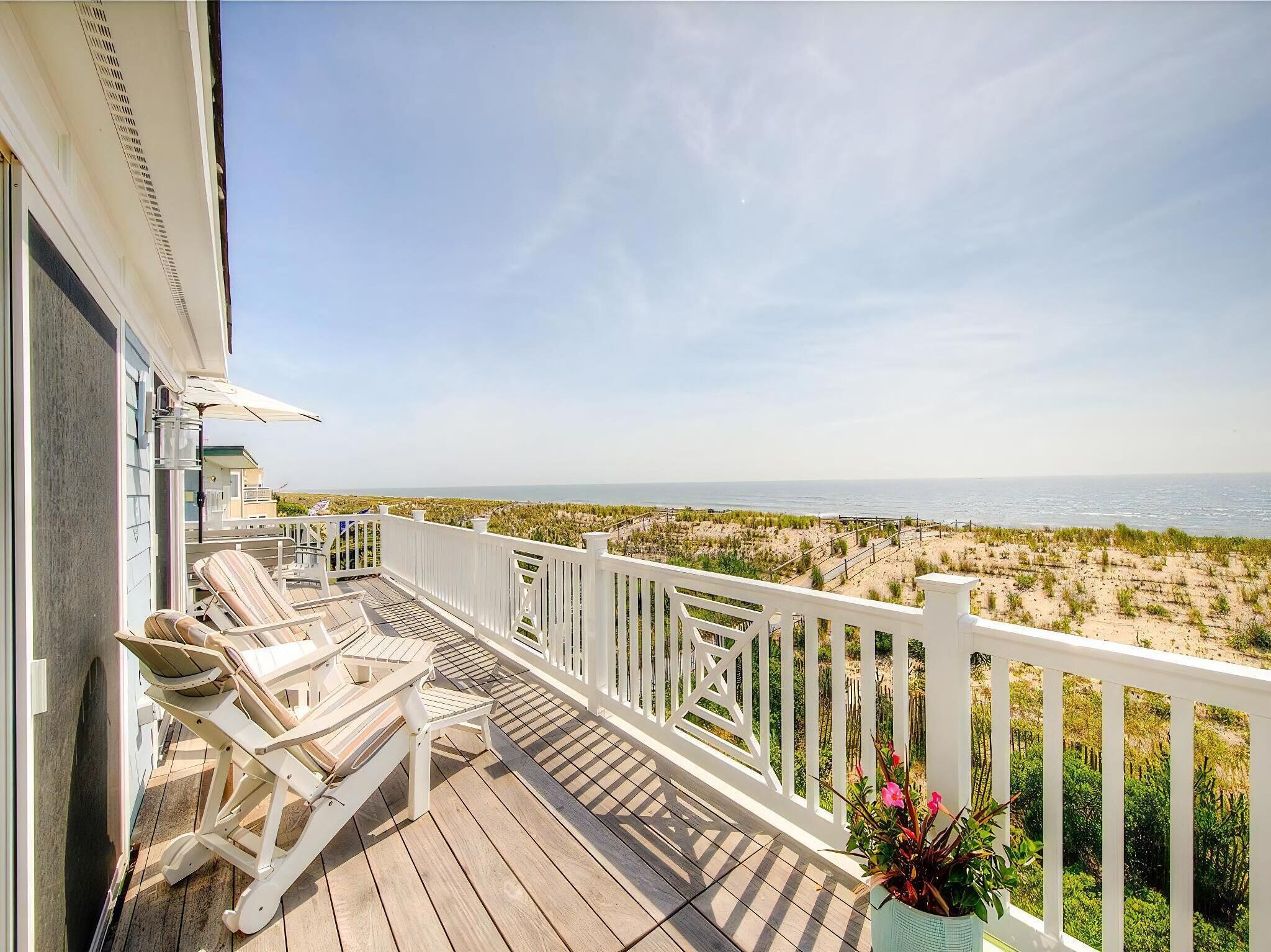 A wooden deck with white railings, overlooking a beach with dunes and the ocean.