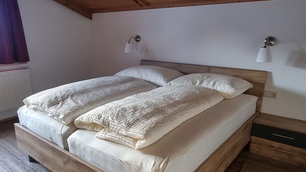 Een tweepersoonsbed met houten frame en witte beddengoed, een houten plafond en een wandlamp.