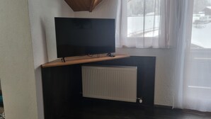 Een kamer met een flatscreen televisie bevestigd aan een houten plank boven een radiator, een raam met dunne gordijnen en een donkere houten vloer.