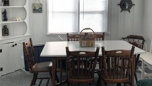 Un coin repas avec une table blanche et des chaises en bois, un ventilateur de plafond et une fenêtre avec des stores.