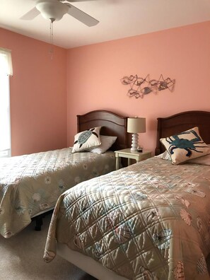 A bedroom with two beds, a ceiling fan, a wall art piece, and a bedside table with a lamp.