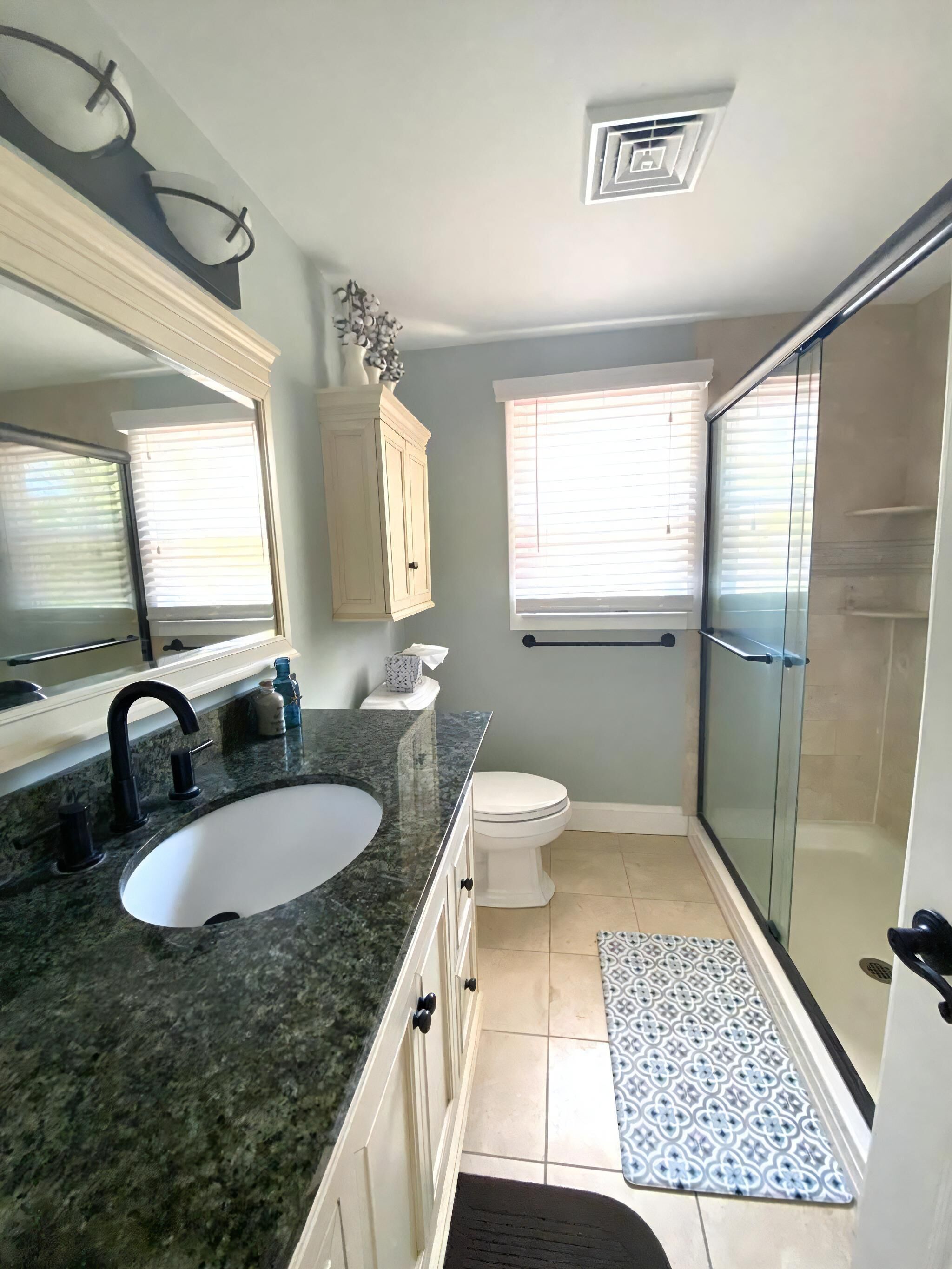 Une salle de bain avec un lavabo à vasque en granit, des toilettes blanches, une douche en verre et un tapis à motifs.
