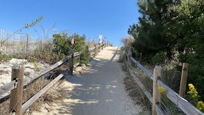 A sandy path with wooden railings, leading to a beach area with vegetation and flags. - OCEANFRONT 1st Floor Duplex (Beach Haven)