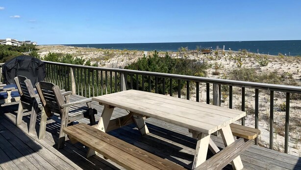 A wooden deck with a picnic table and chairs, overlooking a beach with dunes and the ocean.