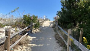 A sandy path with wooden railings, leading to a beach area with vegetation and flags.