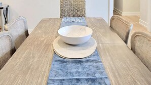 A dining area with a wooden table, a bowl, woven chairs, a hanging light fixture, and a wall art piece. - Ocean view vacation rental on LBI (Ship Bottom)