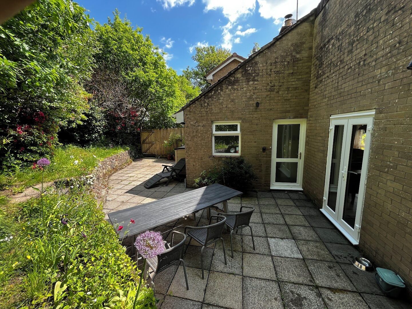 A patio with a table and chairs, a small garden, and a brick building with a door and windows.