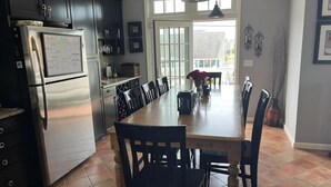 A kitchen with dark cabinets, a stainless steel refrigerator, a dining table with chairs, and a view of the outside through large windows. - Beautiful Raised Ranch with Views of the Bay (Surf City)