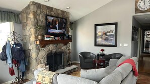 A living room with a stone fireplace, a grey sofa, a television mounted above the fireplace, and a framed picture on the wall. - Beautiful Raised Ranch with Views of the Bay (Surf City)