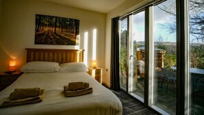 Ein Schlafzimmer mit einem großen Bett, zwei Nachttischlampen, einem Gemälde an der Wand und Blick durch eine Glastür in einen Außenbereich.