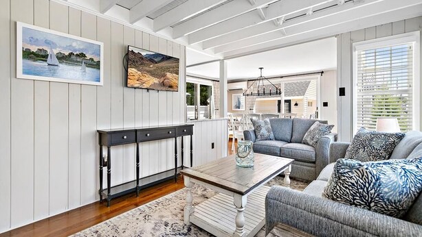 Ein geräumiges Wohnzimmer mit einem Holztisch, einem grauen Sofa und einem gemusterten Teppich. An der Wand hängen zwei Bilder und im Hintergrund befindet sich ein Essbereich.