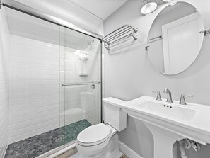 Ein modernes Badezimmer mit einer Glaskabine zur Dusche, weißen Fliesen, einem runden Spiegel und einem weißen Waschbecken mit zwei Wasserhähnen.