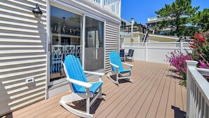 Eine Terrasse mit zwei blauen Adirondack-Stühlen, einer Glastür, die zu einem Raum führt, und einem weißen Geländer.
