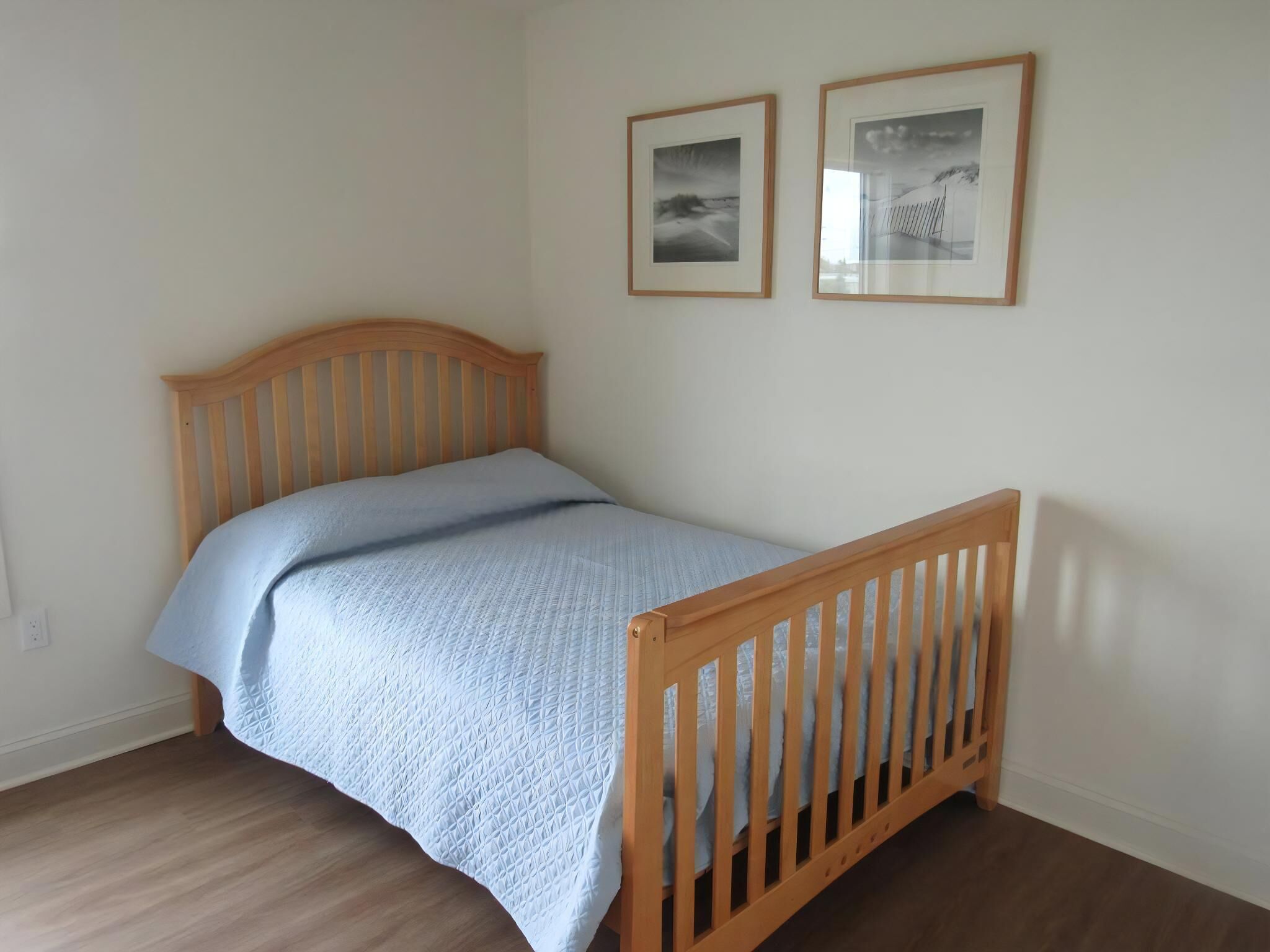 A single bed with a wooden headboard and footboard, a light blue bedspread, and two framed pictures on the wall.