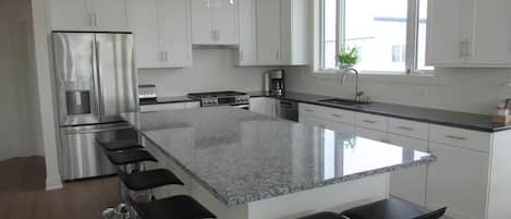 A modern kitchen with white cabinets, a stainless steel refrigerator, a granite countertop, and a large window.
