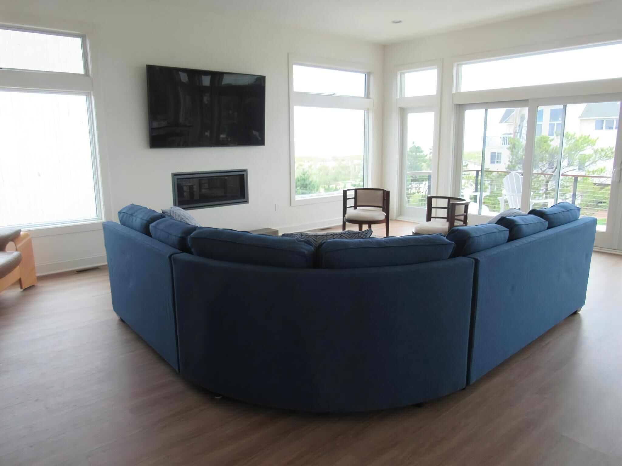 Een moderne woonkamer met een grote blauwe hoekbank, een flatscreen-televisie aan de muur en grote ramen met uitzicht op de natuur.