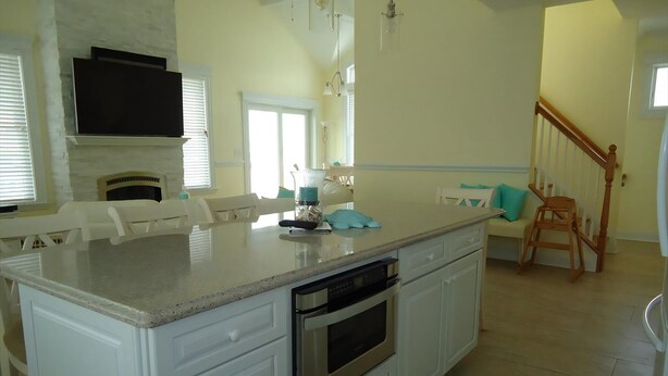 Una cocina con gabinetes blancos, horno empotrado, encimera de granito y una zona de comedor con una silla blanca y un cojín verde azulado.