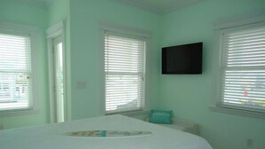 Un dormitorio con cama, persianas blancas, un televisor en la pared y ventana con vistas.