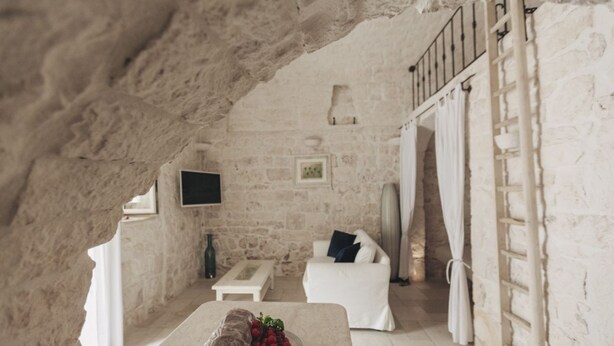 A rustic stone interior with a white sofa, a small table with fruit, and a TV mounted on the wall. - Casetta Garibaldi – Studio apartment in the heart of nineteenth-century Ostuni (Ostuni)