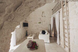 Interior rústico de pedra com um sofá branco, uma pequena mesa com frutas e uma TV fixada na parede.