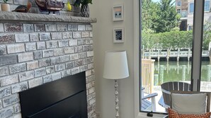 A living room with a brick fireplace, a large window with a view of a body of water, a sofa with patterned cushions, and a small table with a lamp. - 4 bedroom accommodation in Loveladies (Loveladies)