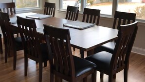 A dining room with a rectangular table and six chairs, large windows with a view of trees and buildings, and wooden flooring.