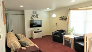 A living room with a sofa, a TV on a stand, a black leather chair, a small table, and a window with curtains.