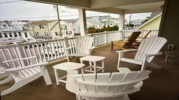 A covered porch with white Adirondack chairs, a small table, and a view of houses in the background. - 220 12th St Associates, LLC (Beach Haven)