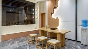 A modern interior with a wooden dining table, two stools, a large window with lights, and a water dispenser. - Yin E Hotel (Emeishan City)