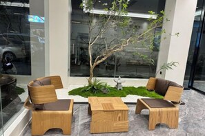 Una zona de estar con mobiliario de madera y un pequeño árbol, ubicada frente a un edificio con fachada de vidrio.