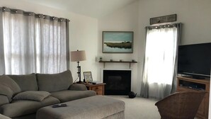 A living room with a sofa, ottoman, TV, fireplace, and a painting on the wall.