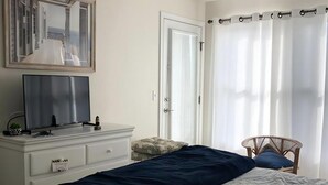 A bedroom with a bed, a ceiling fan, a television on a dresser, a door, and a window with curtains.