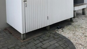 A white storage shed with a door and a small window, situated on a paved area with gravel.