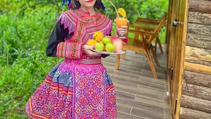 A person in traditional attire holding a plate with fruits and a drink, standing in a wooden structure with a thatched roof, surrounded by greenery. - Bac Ha Lodge Reatreat (Bac Ha)