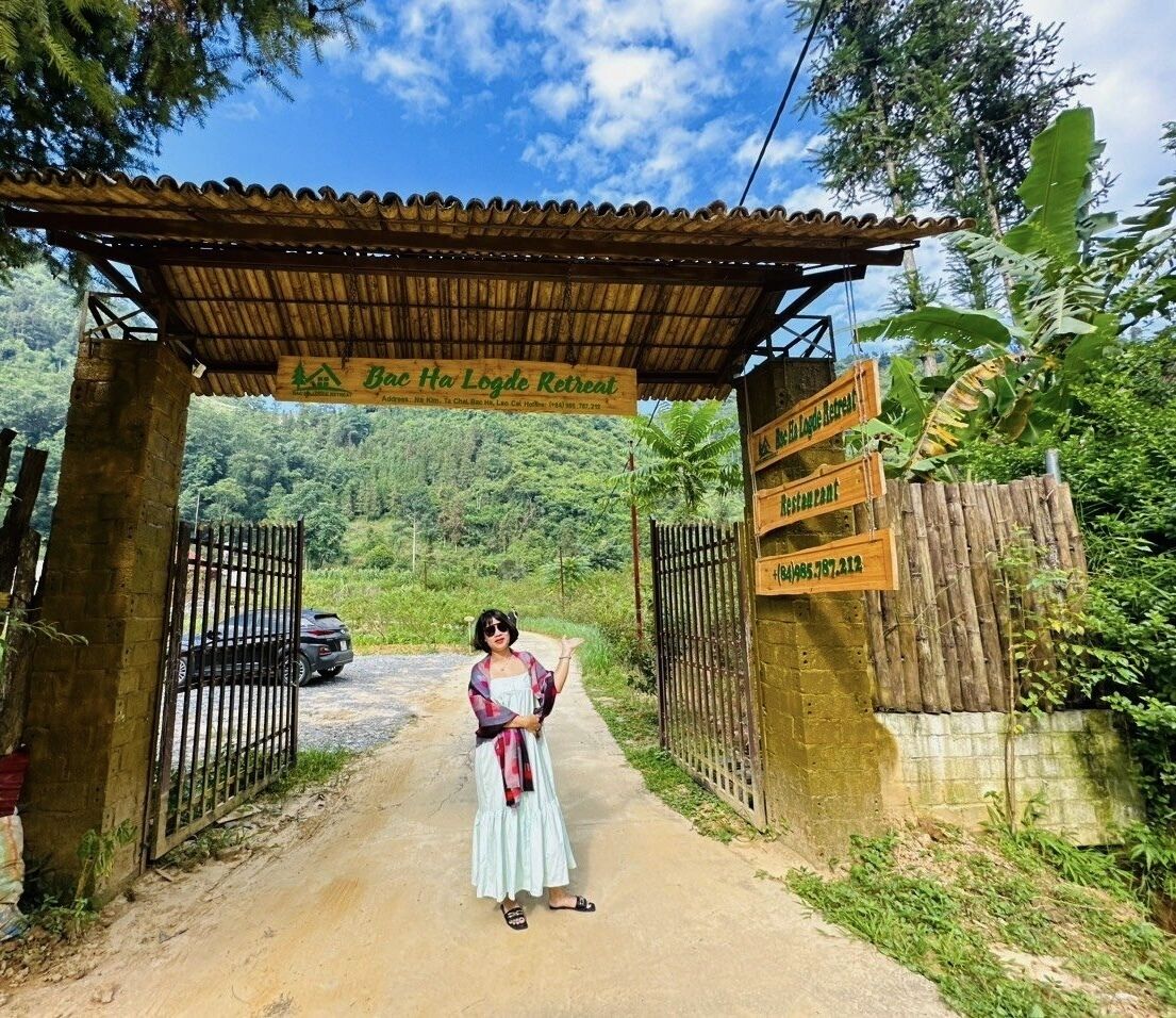 A rustic retreat entrance with a wooden signboard, a metal gate, and a paved path leading into a lush green area.