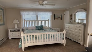 A bedroom with a white bed, a dresser, a nightstand, a ceiling fan, and a window with blinds.