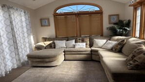 A living room with a sectional sofa, a small ottoman, a large window with wooden shutters, and a potted plant.