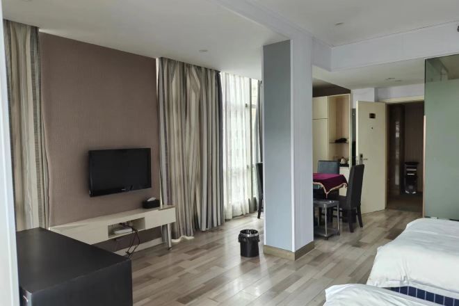 Habitación de hotel con una cama, un televisor sobre un soporte, un escritorio con una silla y un área de comedor visible a través de una puerta abierta.
