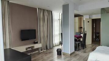 Habitación de hotel con una cama, un televisor sobre un soporte, un escritorio con una silla y un área de comedor visible a través de una puerta abierta.