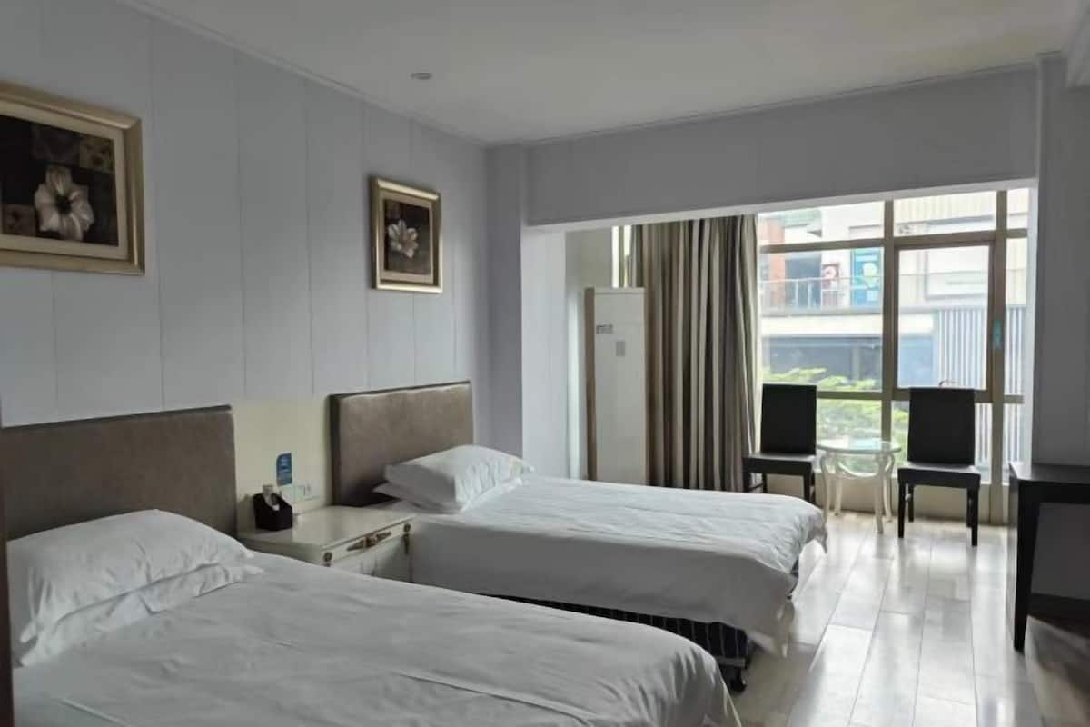 A hotel room with two beds, a small table, and a chair. There is a window with curtains, and a balcony is visible through the window.