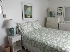 Een slaapkamer met een bed, nachtkastje, dressoir, lamp, een ingelijste foto aan de muur en een prullenbak.