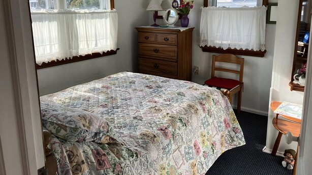Un dormitorio con cama, cómoda, silla, espejo y dos ventanas con cortinas.
