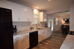 A modern kitchen with white cabinetry, a dishwasher, a coffee maker, and a dining area with a table and chairs. - Beautiful 4 Bedroom in quiet neighborhood! (Bedford)